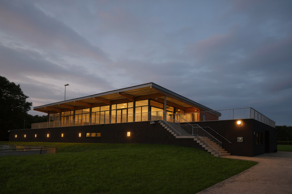 Clubgebouw NWC Asten op sportpark ’t Root in de avond, met verlichte kantine en zicht op de velden. Ontworpen door VDLP architecten. Gebouwd door bouwbedrijf van Gerven.