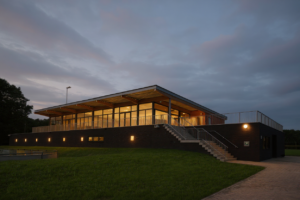 Clubgebouw NWC Asten op sportpark ’t Root in de avond, met verlichte kantine en zicht op de velden. Ontworpen door VDLP architecten. Gebouwd door bouwbedrijf van Gerven.